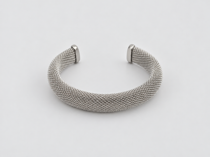 Pulsera Laura, brazalete abierto plateado de malla metálica entrelazada con extremos lisos, sobre fondo gris claro.
